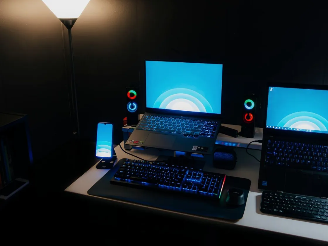 Een donkere werkplek met twee laptops, een verlicht toetsenbord en een lamp